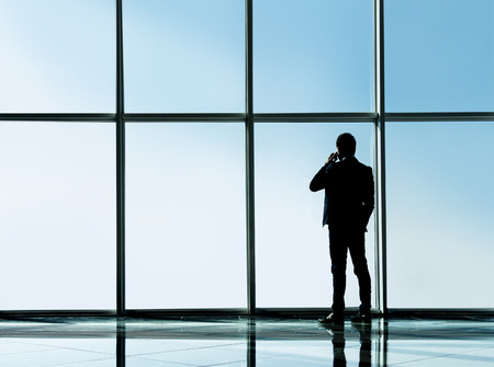Silhouette back view of young businessman is looking out of a panoramic window and talking by phone.の写真素材