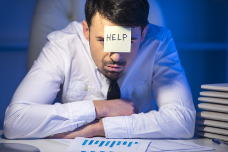 Frustrated young businessman in his office with adhesive note on his forehead. He needs help.の写真素材