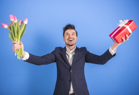 Handsome young man is holding a gift and a flower while standing against blue background.の写真素材