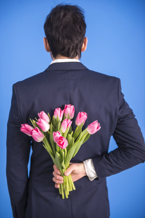 Man is holding bouquet of flowers behind his back isolated on blue background.の写真素材