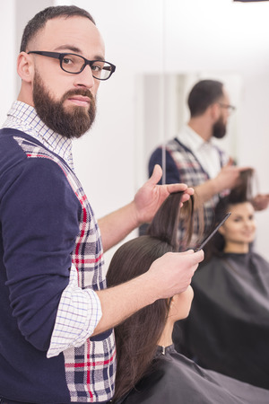 Handsome hairdresser making a haircut for a beautiful brunette girl and looking at camera.の写真素材