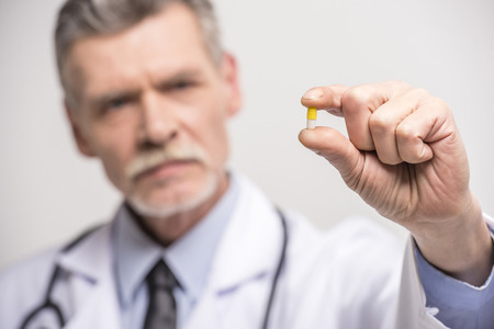 Senior male doctor holding pills on grey background. Focus on pill.の写真素材
