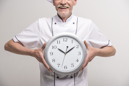 Close-up. Time to dinner. Senior male chief cook in uniform holding a clock on grey background.の写真素材