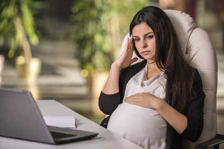 Pregnant adult businesswoman working with laptop looking stressed in office.の写真素材