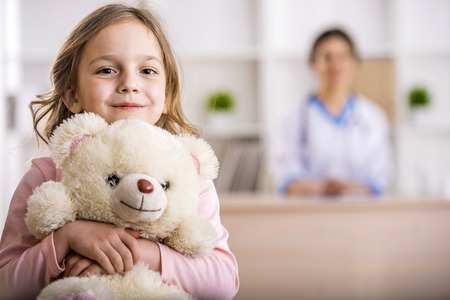 Little girl with teddy bear is looking at the camera. Female doctor on background. Little girl with teddy bear is looking at the camera. Female doctor on background.