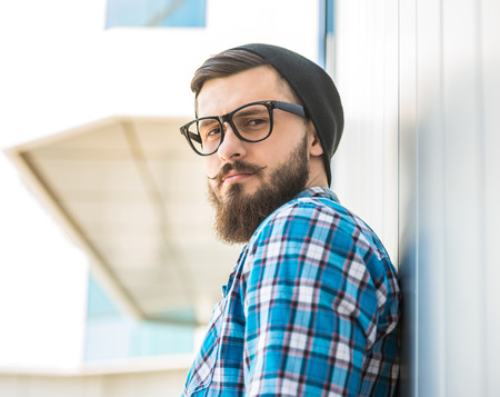 Young trendy man is standing outdoor in hat and glasses and looking at the camera.の写真素材