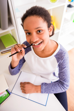 Young pretty smiling mulatto schoolgirl sitting at the table and holding a pencil in hand on colorful background.の写真素材
