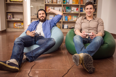 Two young handsome guys sitting on poufs and playing video games together. Leisure time.