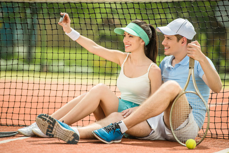 Smiling young couple sitting on tennis court, holding tennis racket and making selfie.の写真素材