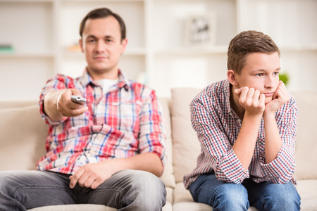 Boy sitting at sofa and get bored while his father watching television.の写真素材