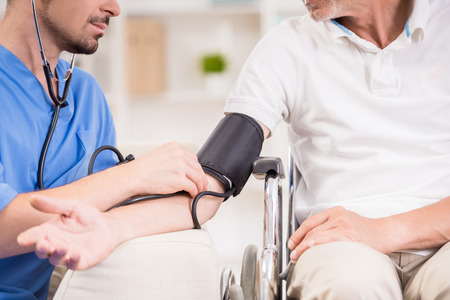 Male doctor measuring blood pressure to older patient sitting at wheelchair.