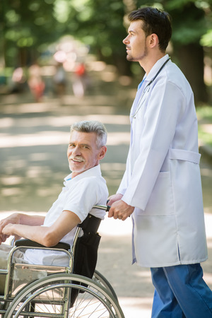 Male nurse walking with senior patient in wheelchair in garden near hospital. Side view.の写真素材