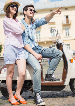 Beautiful young couple dressed casual sitting on scooter together, man pointing away.の写真素材