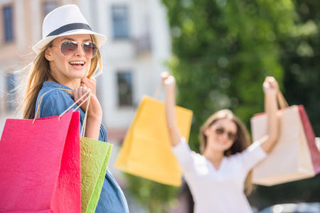 Beautiful young woman with shopping bags looking at camera and smiling while her friend standing on background.の写真素材