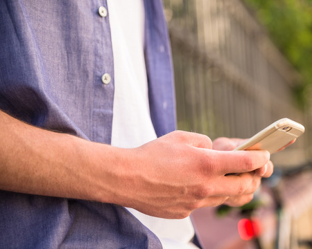 Young man holding his phone, standing near the fence outdoors. Close-up.の写真素材