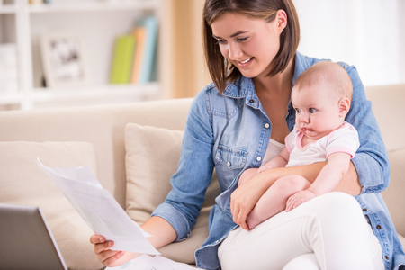 Young woman is working from home, holding baby girl on lap.の写真素材