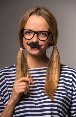 Beautiful young woman with two tails is holding fake mustache on her face and smiling.の写真素材
