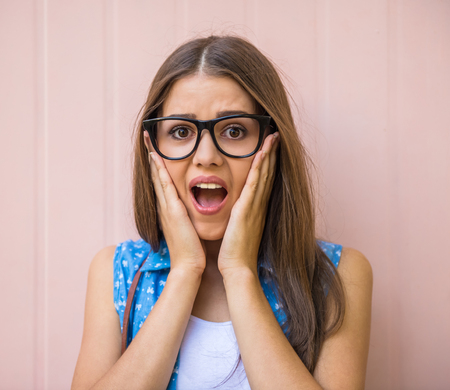Surprised young woman in glasses is holding hands on face.