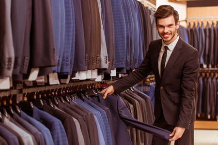 Modern young handsome businessman smiling, looking and choosing classical suit in the suit shopの写真素材