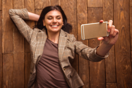 Top view of beautiful girl in casual clothes making selfie and smiling while lying on wooden floor