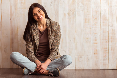 Beautiful girl in casual clothes looking in camera and smiling while sitting against wooden backgroundの写真素材