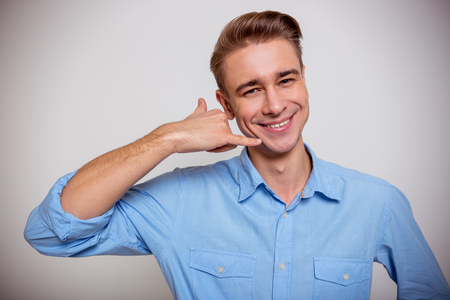Portrait of attractive young blond businessman in blue shirt smiling and imitating phone with his hand, standing against gray backgroundの写真素材