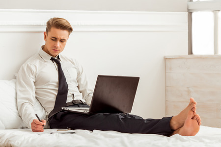Attractive young blond businessman in white classical shirt and dark tie making notes and using laptop while sitting on bedの写真素材