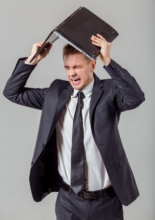 Portrait of attractive young blond businessman in classical suit screaming and holding laptop overhead, standing against gray backgroundの写真素材