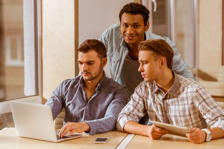 Three young handsome businessmen in casual clothes smiling, discussing ideas, using laptop and tablet while working in officeの写真素材