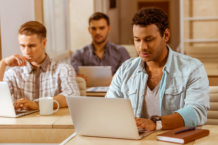 Young handsome mulatto businessman in casual clothes using laptop, in the background two other businessmen working in office, using laptop and tabletの写真素材