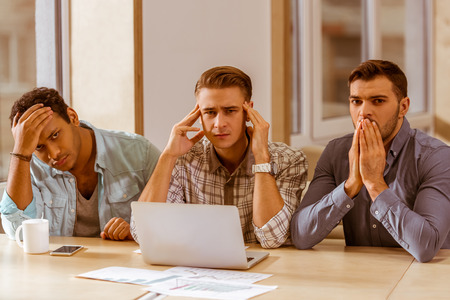 Three upset young handsome businessmen in casual clothes using laptop while sitting in office, looking in cameraの写真素材