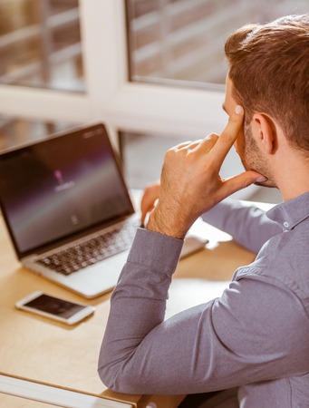 Young handsome dark-haired businessman in casual clothes using laptop while working in office, backの写真素材