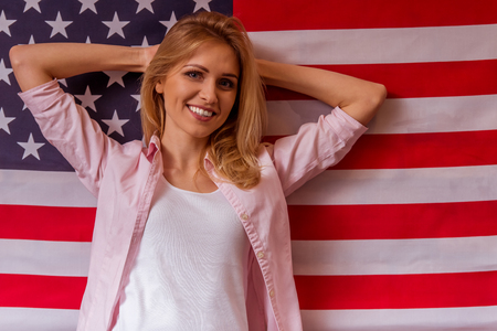 Beautiful young blonde girl in casual clothes posing, smiling and looking in camera, hands behind head, standing against American flagの写真素材