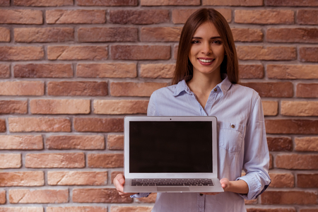 Beautiful young dark-haired girl in casual clothes posing, smiling and showing laptop, standing against brick wallの写真素材