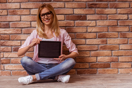 Beautiful young blonde girl in casual clothes posing, smiling and showing tablet, sitting cross-legged against brick wallの写真素材