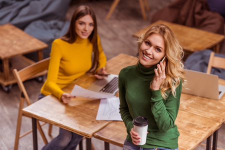Top view of beautiful young blonde girl in casual clothes holding cup and talking on the phone, standing in cafe, in the background dark-haired girl using laptopの写真素材