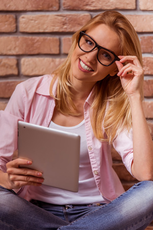 Beautiful young blonde girl in casual clothes posing, smiling and looking in camera, sitting cross-legged against brick wall with tabletの写真素材