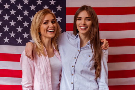 Two beautiful young girls in casual clothes posing, smiling and looking in camera, standing against American flagの写真素材