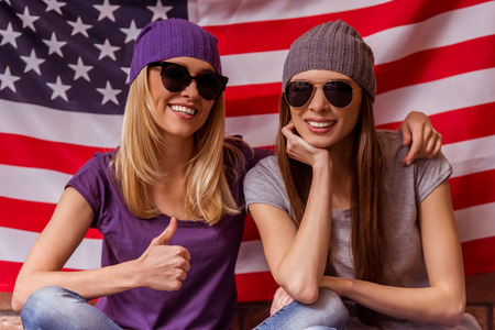 Two beautiful young girls in casual clothes and sunglasses posing, smiling and showing OK , sitting cross-legged against American flagの写真素材