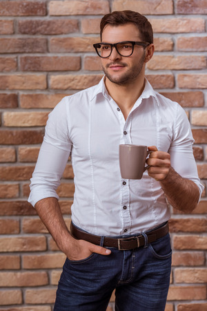 Portrait of attractive young businessman in white shirt, jeans and eyeglasses holding a cup and looking away while standing against brick wallの写真素材