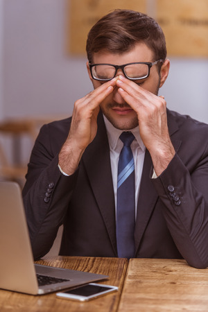 Attractive young businessman in classical suit and eyeglasses is tired after working in the cafeの写真素材