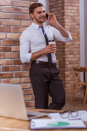 Attractive young businessman in white classical shirt talking on the mobile phone, holding a cup and smiling while working in the cafeの写真素材