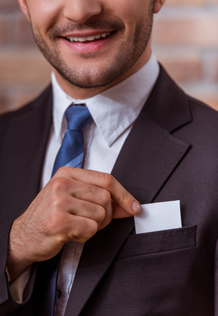 Portrait of attractive young businessman in classical suit holding a white card and smiling, standing against brick wall, close-upの写真素材