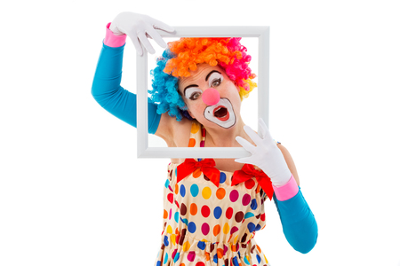 Portrait of a funny playful female clown in colorful wig holding a white frame, looking at camera and showing emotions, isolated on a white backgroundの写真素材