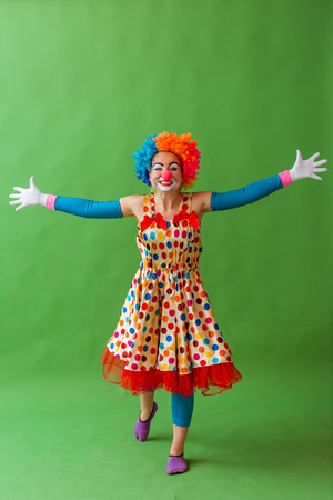 Funny playful female clown in colorful wig keeping hands apart, looking at camera and smiling, standing on a green backgroundの写真素材