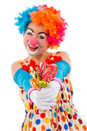 Portrait of a funny playful female clown in colorful wig holding lollipops, looking at camera and smiling, isolated on a white background, focus on handsの写真素材