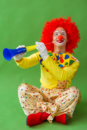 Funny playful clown in red wig holding a blowing horn, looking at camera and smiling, sitting on a green backgroundの写真素材