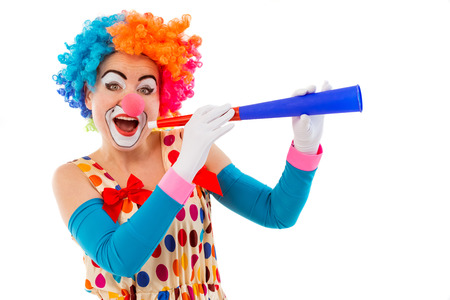 Portrait of a funny playful female clown in colorful wig blowing horn, looking at camera and smiling, isolated on a white backgroundの写真素材