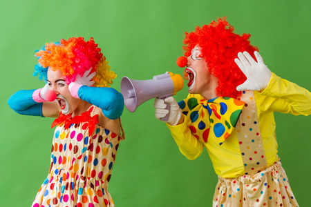 Two funny playful clowns standing on greenc background. Woman covering her ears while man screaming...