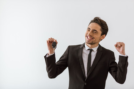 Happy handsome young businessman in classic suit smiling, looking away, holding keys and showing fist, on white backgroundの写真素材